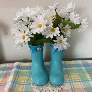 May include: A pair of light blue decorative rain boots filled with white daisy flowers. The boots have a polka dot ribbon and bow detail. The arrangement sits on a white lace doily and a plaid tablecloth.