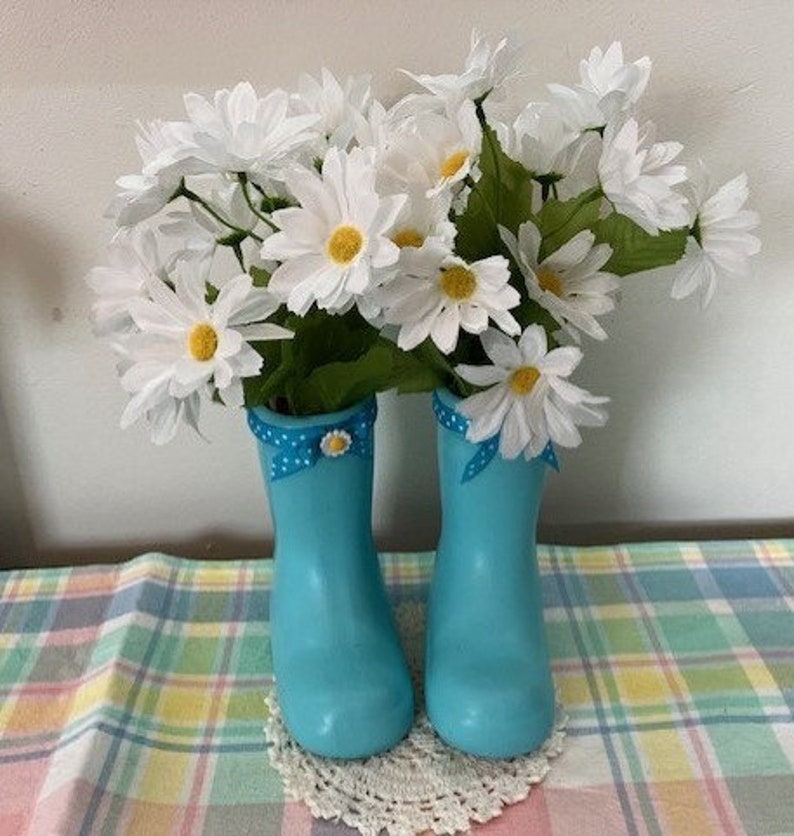 May include: A pair of light blue decorative rain boots filled with white daisy flowers. The boots have a polka dot ribbon and bow detail. The arrangement sits on a white lace doily and a plaid tablecloth.