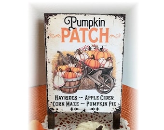 May include: A rectangular wooden sign with the words "Pumpkin Patch" in black and orange. The sign features a wheelbarrow and a wooden barrel filled with pumpkins. The bottom of the sign reads "Hayrides ~ Apple Cider ~ Corn Maze ~ Pumpkin Pie."