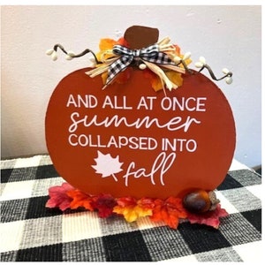 May include: An orange wooden pumpkin-shaped sign with the text "AND ALL AT ONCE summer COLLAPSED INTO fall". It is decorated with autumn leaves, a black and white bow, and an acorn.