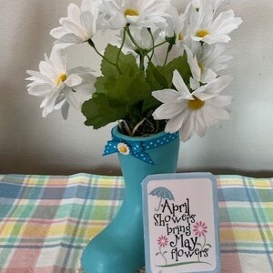 May include: A decorative arrangement featuring white daisy-like flowers in a light blue rain boot vase. A small sign with the text "April Showers bring May flowers" sits in front of the boot. The arrangement is on a plaid surface.