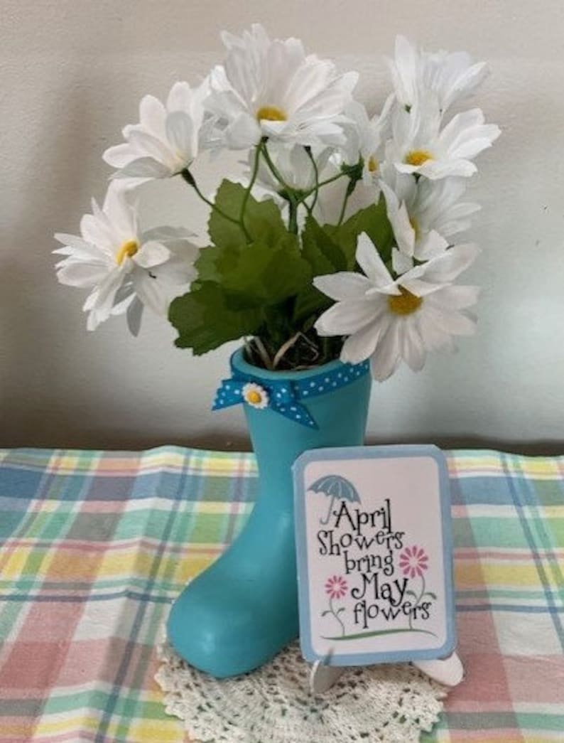 May include: A decorative arrangement featuring white daisy-like flowers in a light blue rain boot vase. A small sign with the text "April Showers bring May flowers" sits in front of the boot. The arrangement is on a plaid surface.