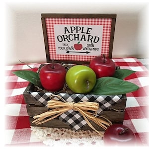 May include: A wooden crate containing red and green apples and green leaves. Above the crate is a sign that reads "Apple Orchard" and "Pick Your Own". The crate is lined with a black and white checked fabric and tied with raffia.