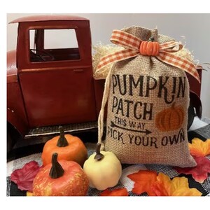 May include: A burlap bag with the text "Pumpkin Patch This Way Pick Your Own" and a pumpkin graphic. The bag is tied with a gingham ribbon and topped with a small pumpkin. Decorative pumpkins and fall leaves are in front of a red truck.