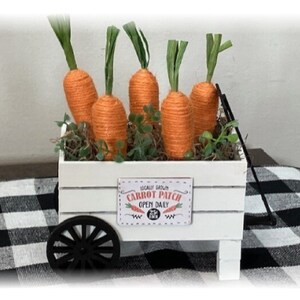May include: A white wooden cart filled with orange twine carrots and greenery. The cart has a black wheel and a sign that reads "Locally Grown Carrot Patch Open Daily". A decorative Easter or spring theme.