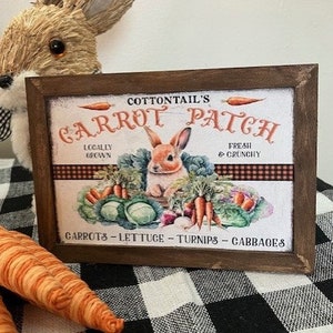 Może przedstawiać: Drewniany znak z brązową ramą i białym tłem. Znak głosi "Cottontail's Carrot Patch" pomarańczowymi literami. Na znaku znajduje się obraz królika i warzyw. Tekst pod obrazem głosi "Locally Grown" i "Fresh & Crunchy". Znak głosi również "Carrots - Lettuce - Turnips - Cabbages".