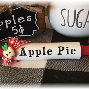 May include: A decorative rolling pin with red handles and the words "Apple Pie" printed on the white barrel. An apple-shaped embellishment with a red and white gingham ribbon is attached. A sign reads "apples 5¢".