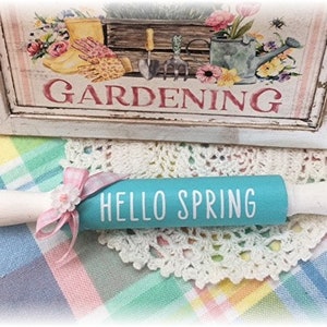 May include: A wooden rolling pin with white handles and a turquoise centre that reads "HELLO SPRING" in white. A pink ribbon and flower accent the pin. A decorative sign with the word "GARDENING" is in the background.