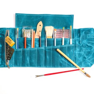 May include: A teal blue canvas roll-up case with multiple pockets for holding art supplies. The case is open and filled with brushes, pencils, and a small eraser.