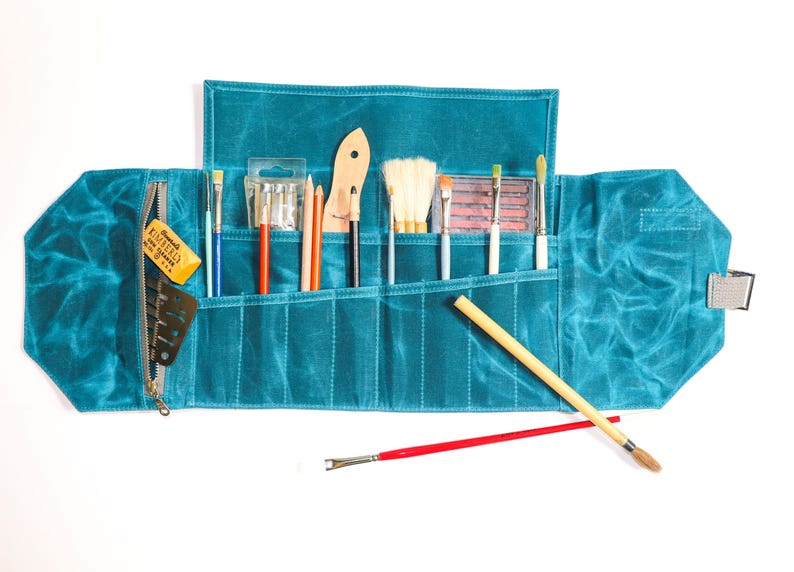 May include: A teal blue canvas roll-up case with multiple pockets for holding art supplies. The case is open and filled with brushes, pencils, and a small eraser.