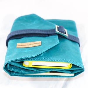 May include: A teal blue canvas roll-up case with a dark blue strap and silver buckle. The case is closed and has a label that reads "nightbloomingjournal".