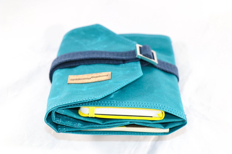 May include: A teal blue canvas roll-up case with a dark blue strap and silver buckle. The case is closed and has a label that reads "nightbloomingjournal".