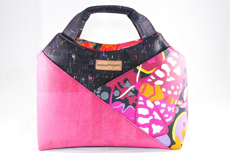 May include: A black, pink, and multi-colored tote bag with a cork bottom and a fabric handle. The bag has a geometric design with a butterfly pattern and the text "Jumping Pineapple" on the front.