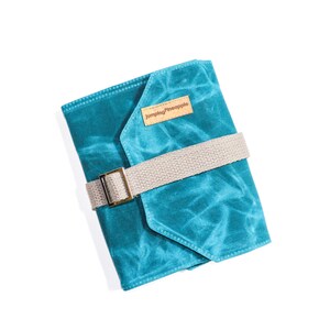 May include: A teal blue canvas notebook cover with a brown leather patch that says "Jumping Pineapple". The cover has a beige strap with a gold buckle.