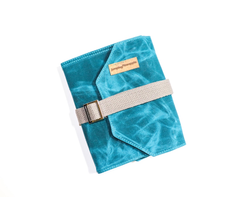 May include: A teal blue canvas notebook cover with a brown leather patch that says "Jumping Pineapple". The cover has a beige strap with a gold buckle.