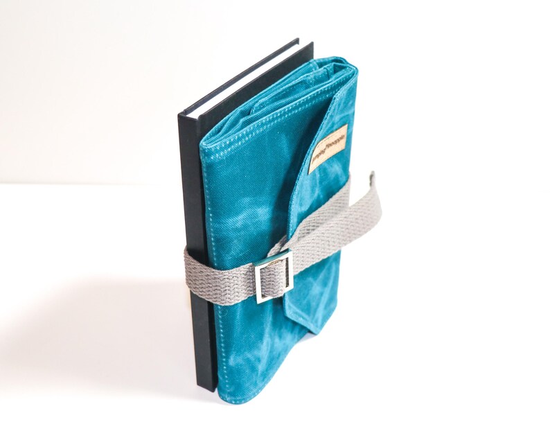 May include: A teal blue canvas notebook with a grey strap and buckle closure. The notebook is wrapped around a black hardback journal.