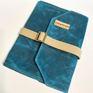 May include: Teal blue canvas journal with a tan strap and buckle closure. The journal has a brown leather patch with the text "Jumping Pineapple" on it.