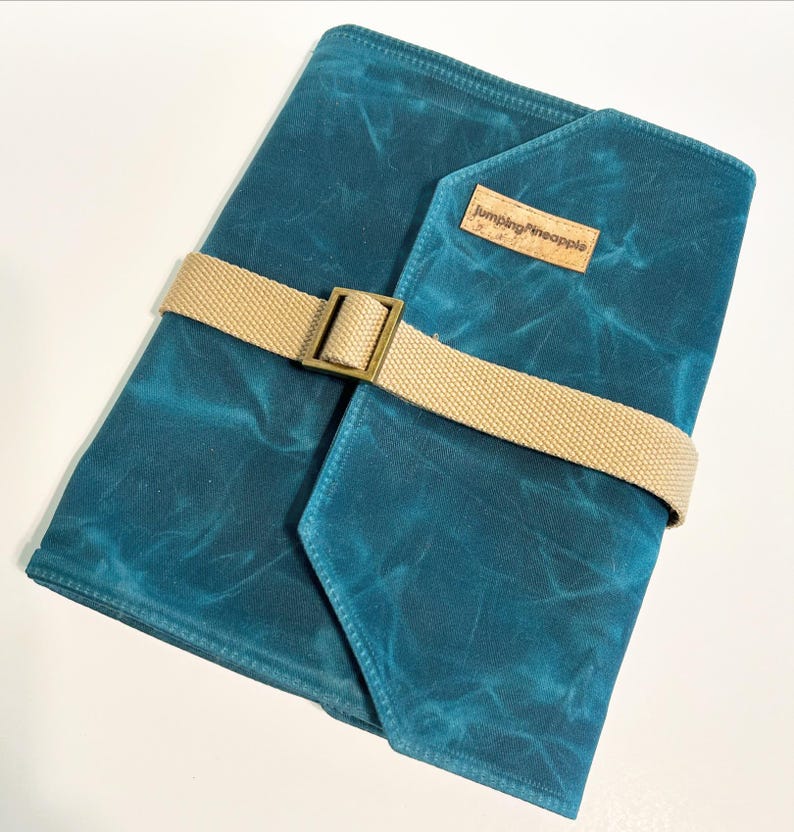 May include: Teal blue canvas journal with a tan strap and buckle closure. The journal has a brown leather patch with the text "Jumping Pineapple" on it.