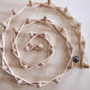 Könnte beinhalten: Ein Murmelbahnspiel aus Holz mit Spiraldesign. Die Bahn besteht aus gewebten, hellfarbigen Holzstreifen mit kleinen Holzstiften. Eine dunkelgraue Murmel liegt am Ende der Bahn. Das Spiel befindet sich auf einer hellbraunen Oberfläche.