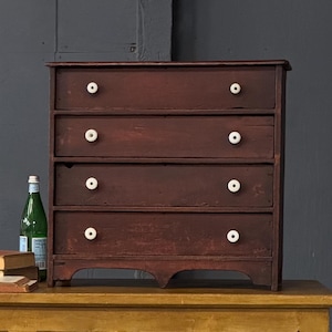 May include: A two-piece antique furniture set. The bottom piece is a mustard-yellow cabinet with three drawers and a paneled front. The top piece is a small, dark red chest of drawers with white knobs. A green bottle and books are on top.