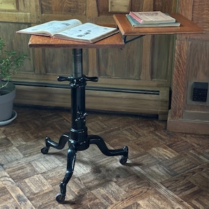 May include: A height-adjustable wooden reading stand with a black cast iron base featuring three legs on casters. The stand has two rectangular wooden surfaces, one holding an open book and the other holding two stacked books, connected by a metal arm.