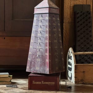 May include: An antique Hohner Harmonicas display case. The burgundy-colored case is pyramid-shaped with a rectangular base. It features text and numbers, with a crank on the side. The base reads "Hohner Harmonicas."