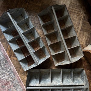 May include: Three vintage metal storage bins with multiple compartments. The bins are a weathered gray color with signs of rust, suggesting age and use. The bins are arranged on a wooden floor.