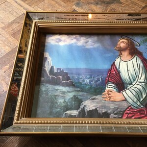 May include: A framed painting of a man in a white and red robe praying with his hands clasped together. The painting is set against a backdrop of a city and a blue sky with clouds.