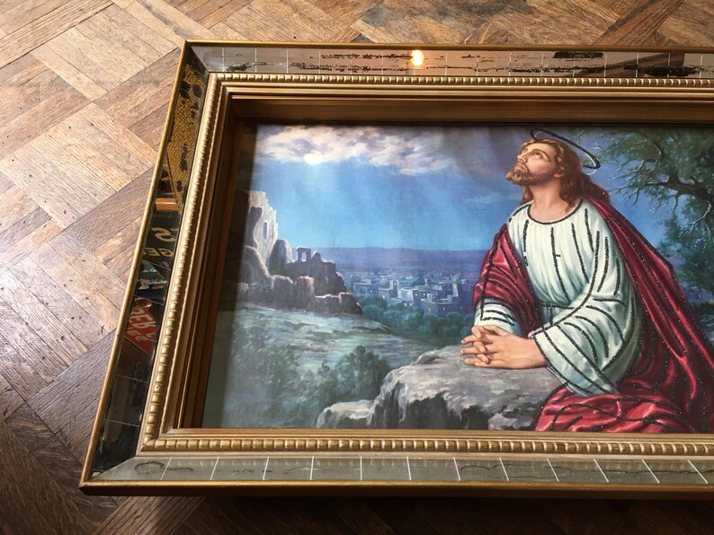 May include: A framed painting of a man in a white and red robe praying with his hands clasped together. The painting is set against a backdrop of a city and a blue sky with clouds.