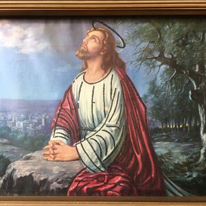 May include: A framed painting of Jesus Christ praying with his hands clasped together. He is wearing a white robe with a red cloak and a halo above his head. The background is a landscape with a city in the distance and trees in the foreground.