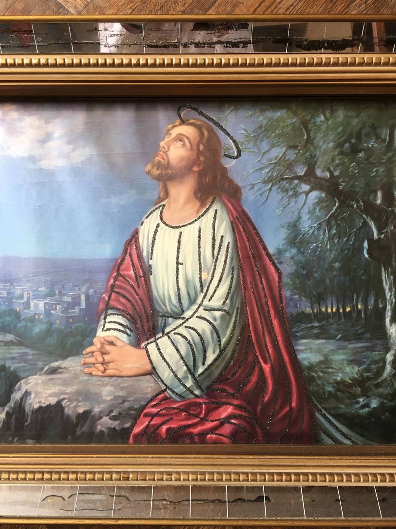 May include: A framed painting of Jesus Christ praying with his hands clasped together. He is wearing a white robe with a red cloak and a halo above his head. The background is a landscape with a city in the distance and trees in the foreground.
