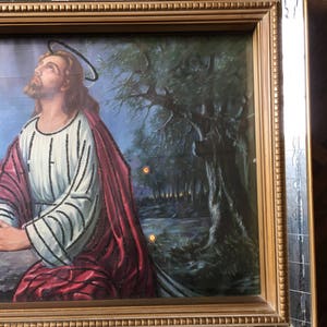 May include: A framed painting of Jesus Christ praying in a forest setting. The painting is in a gold frame with a mirrored border. The painting features a dark blue sky with a halo above Jesus' head. Jesus is wearing a white and red robe with a black beaded design. The forest is depicted in shades of green and brown.