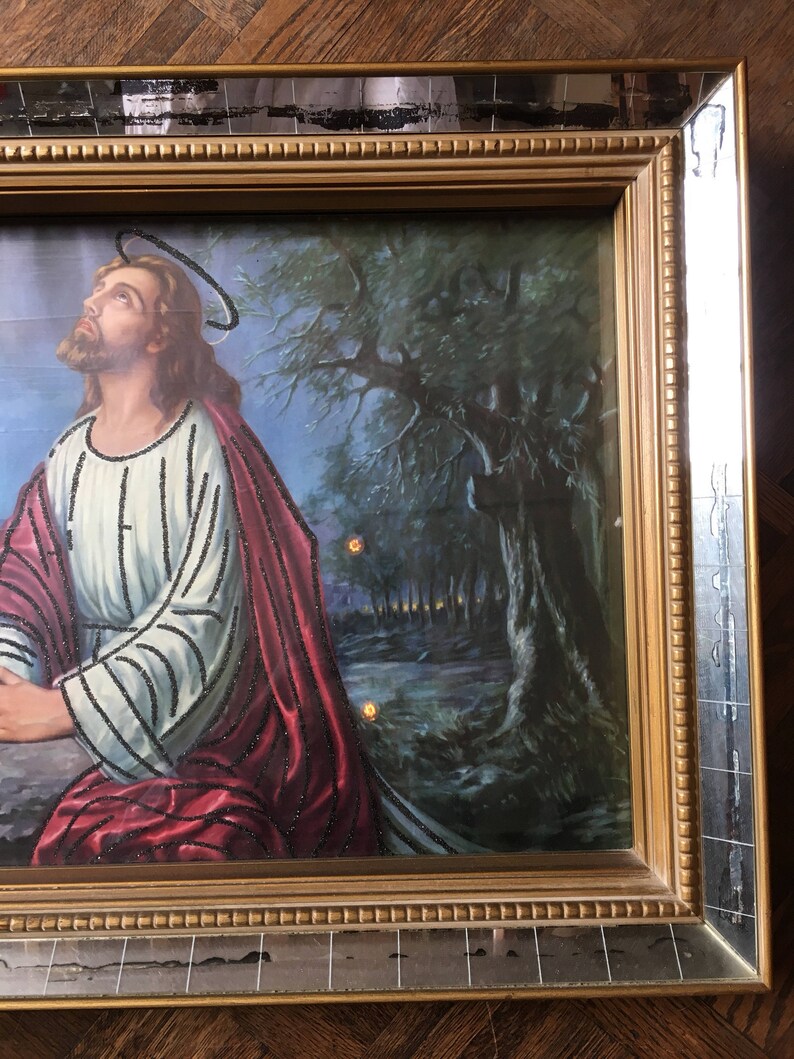 May include: A framed painting of Jesus Christ praying in a forest setting. The painting is in a gold frame with a mirrored border. The painting features a dark blue sky with a halo above Jesus' head. Jesus is wearing a white and red robe with a black beaded design. The forest is depicted in shades of green and brown.