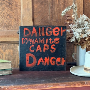 Puede incluir: Un letrero negro con letras rojas que dice "DANGER DYNAMITE CAPS DANGER". El letrero está sobre una superficie de madera con un sombrero y un jarrón de flores secas.