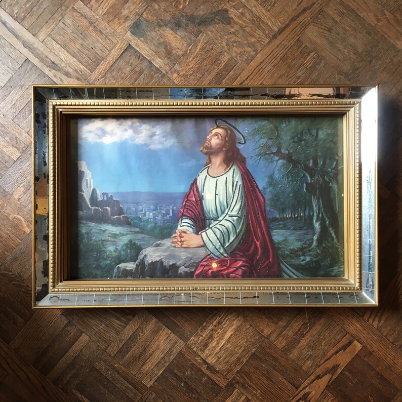 May include: A framed religious print depicting a man in a white robe with a red sash kneeling in prayer. The background shows a city in the distance and a tree in the foreground.
