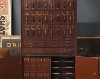 May include: A large, antique, dark red cabinet with numerous drawers and pull handles. The cabinet has a rectangular shape with a flat top and two doors at the bottom. The drawers have label holders with gold trim. The cabinet is a vintage piece of furniture.