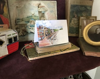 May include: A watercolor painting of a city street scene with colorful buildings, a bridge, and a river. The painting is on a white card with a light blue border.
