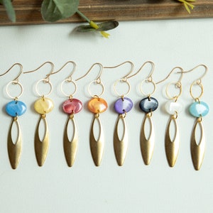 May include: A collection of seven gold-colored earrings. Each earring features a colorful, oval-shaped bead, a gold ring, and a long, gold-colored, teardrop-shaped pendant. The beads are in various colors, including blue, yellow, red, orange, purple, black, and white.