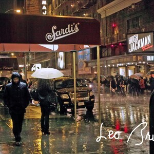Può includere: Una scena notturna piovosa a New York City con un tendone rosso che recita "Sardi's" sopra un SUV nero. Il cartello dell'Helen Hayes Theatre è visibile sullo sfondo. Un cartello per "Hugh Jackman" è visibile sull'edificio a destra.