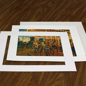 Könnte beinhalten: Eine weiße Passepartout-Karte mit einer kleineren weißen Passepartout-Karte im Inneren, beide mit einer rechteckigen Öffnung. Ein Foto eines Karussells mit Menschen, die auf den Pferden reiten, befindet sich in der Öffnung.