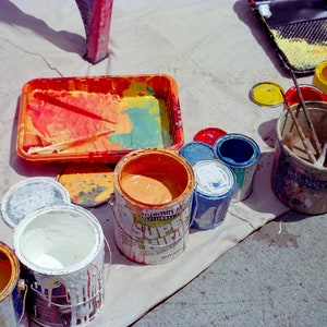 May include: A variety of paint cans and trays with different colors of paint, including red, blue, green, yellow, and white. The paint is in various stages of being used, with some cans nearly empty and others full. There is also a bucket with paint brushes and a paint roller.