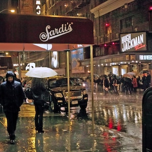 Può includere: Una scena notturna piovosa a New York City con un tendone rosso sopra l'ingresso del ristorante "Sardi's". Il cartello dello spettacolo di Broadway "Hugh Jackman" è visibile sullo sfondo.