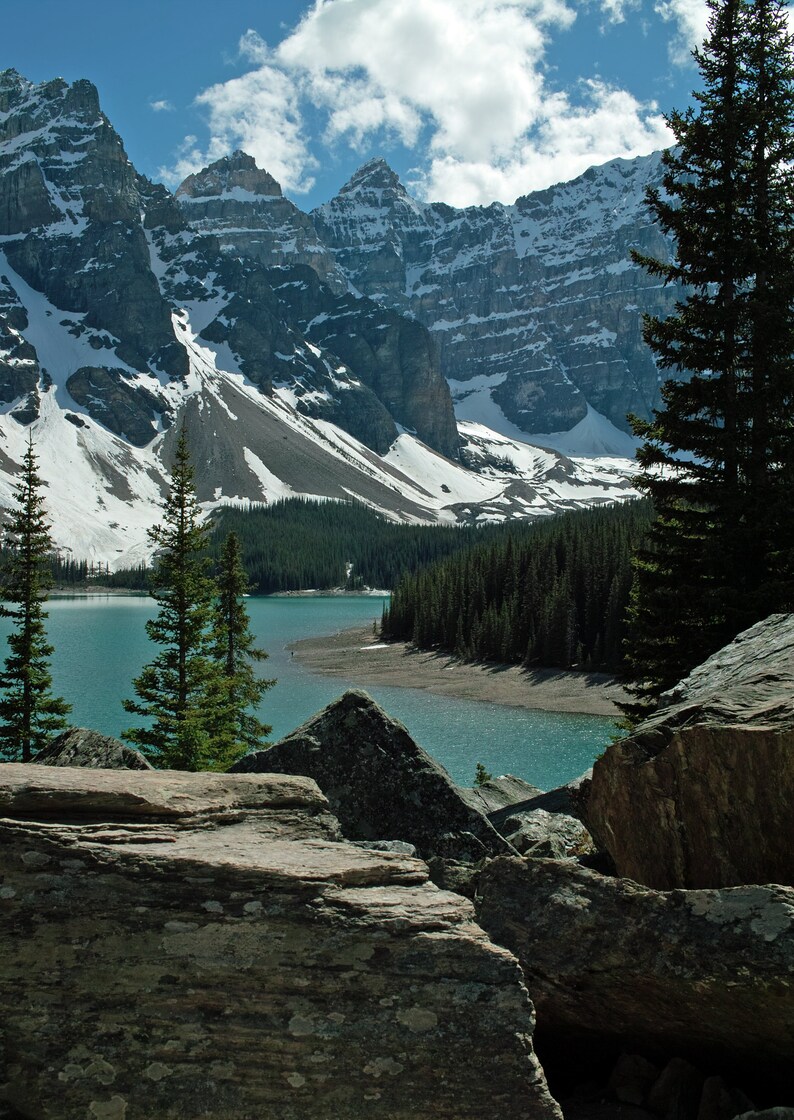 May include: A scenic view of a turquoise lake surrounded by snow-capped mountains and evergreen trees. The lake is nestled in a valley with rocky outcroppings.