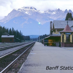 May include: A vintage photograph of a train station in Banff, Canada. The station is a wooden building with a red roof and a sign that reads "Banff Station by Leo Bruce". There are mountains in the background and a train track in the foreground.
