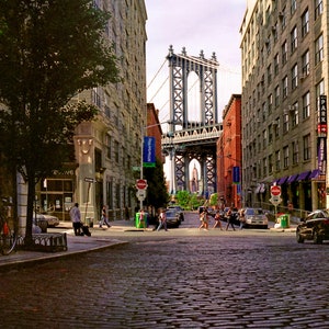May include: A view down a cobblestone street in New York City with the Manhattan Bridge in the background. The street is lined with brick buildings and there are people walking on the sidewalk.