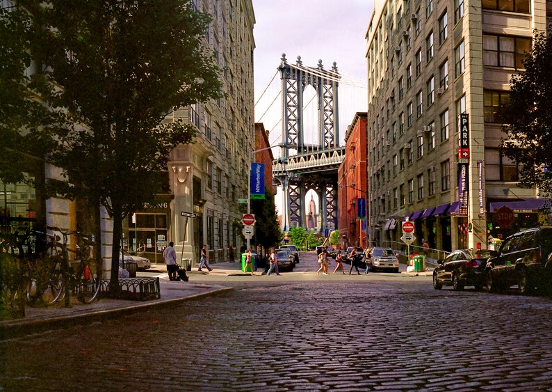 May include: A view down a cobblestone street in New York City with the Manhattan Bridge in the background. The street is lined with brick buildings and there are people walking on the sidewalk.