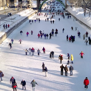 May include: A large group of people ice skating on a frozen canal in the winter. The canal is surrounded by snow-covered buildings and trees. The people are wearing winter clothing and are enjoying the winter weather.