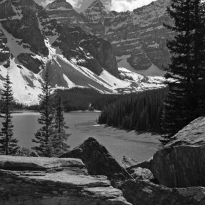 May include: A black and white photograph of a mountain lake with snow-capped mountains in the background. The lake is surrounded by trees and rocks.