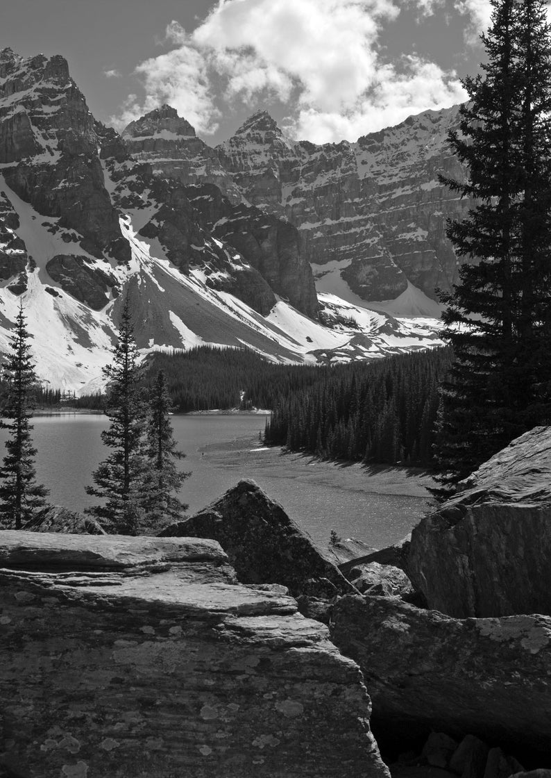 May include: A black and white photograph of a mountain lake with snow-capped mountains in the background. The lake is surrounded by trees and rocks.