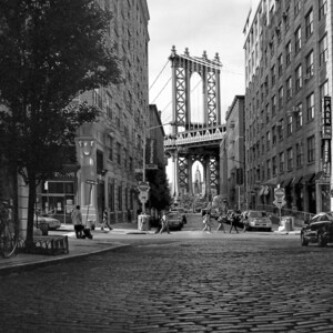 May include: A black and white photo of a street in New York City with the Manhattan Bridge in the background. The street is lined with brick buildings and there are people walking on the sidewalk.